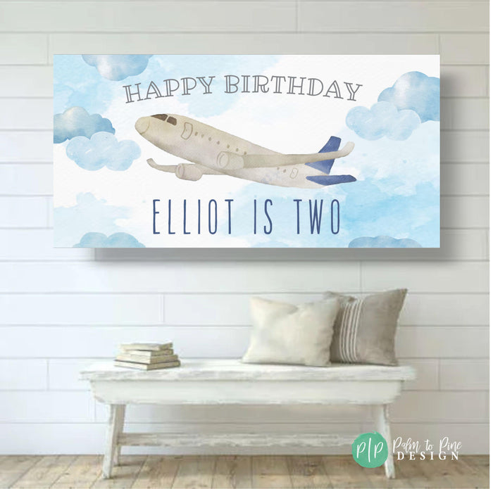 A blue and white airplane flying in a blue sky with white clouds. The text 'Happy Birthday' is at the top of the banner. The text 'Elliot is Two' is at the bottom of the banner.