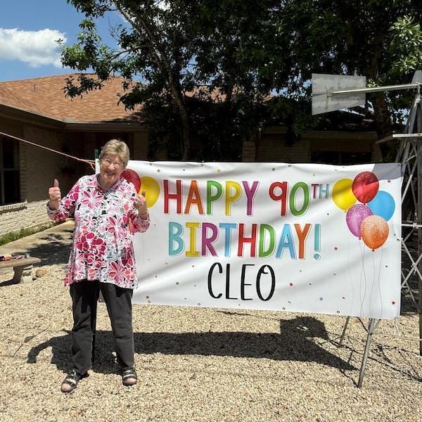 A woman standing in front of a banner that says "Happy 90th Birthday! Cleo" is smiling and giving a thumbs up.