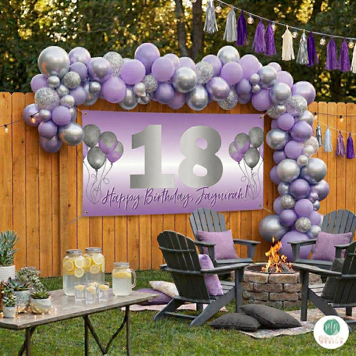 Teen girls purple and silver 18th birthday party set in backyard. Features a personalized vinyl banner hung from the fence and large balloon garland with backyard firepit setting