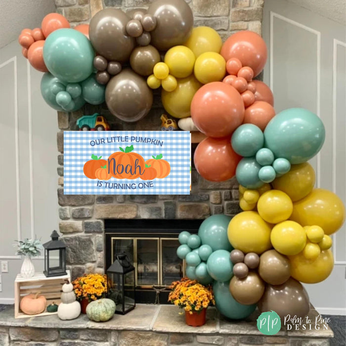 a festive arrangement of colorful balloons in the shape of a pumpkin, with a banner that reads "Our little pumpkin Noah is turning one" displayed above it. The arrangement is set against a backdrop of a stone fireplace, and there are some potted plants and other decorative items in the room.