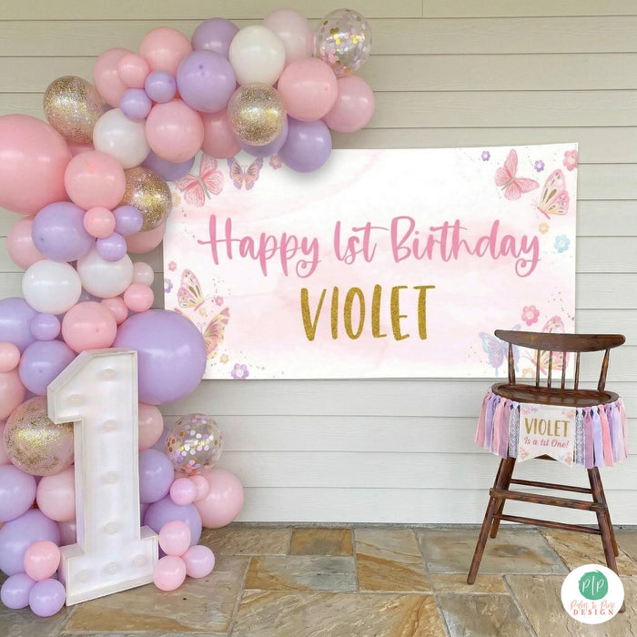 Personalized pink butterfly birthday banner in soft pink and purple watercolor style displayed at a first birthday high chair cake smash photoshoot