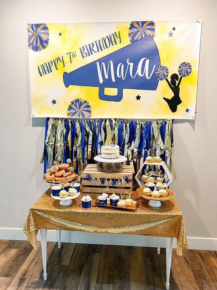 A cheer themed birthday party setup with a table filled with various desserts, including cupcakes, donuts, and a cake, all placed on a gold tablecloth. Above the table, there is a banner that reads "Happy 7th Birthday Mara" and features a cheerleader silhouette and stars. The background is decorated with blue and gold streamers, creating a festive atmosphere.