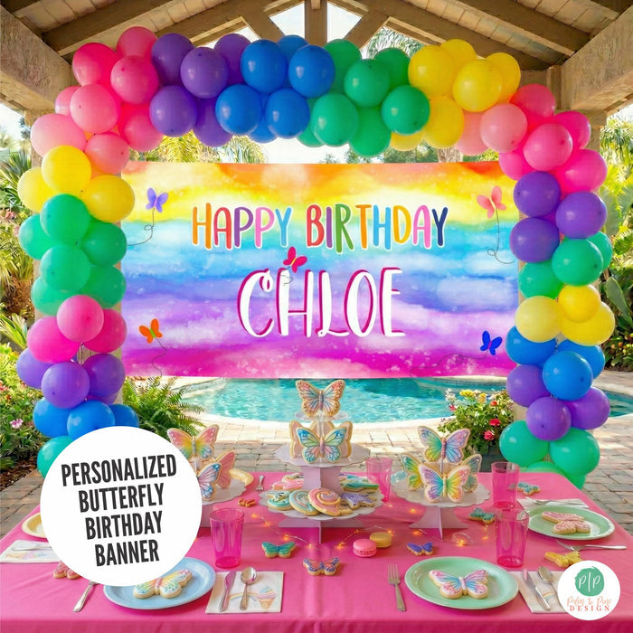 personalized butterfly birthday banner with vibrant colors and whimsical butterfly accents displayed at outdoor girls birthday party with rainbow colored balloon garland and cheerful table set up