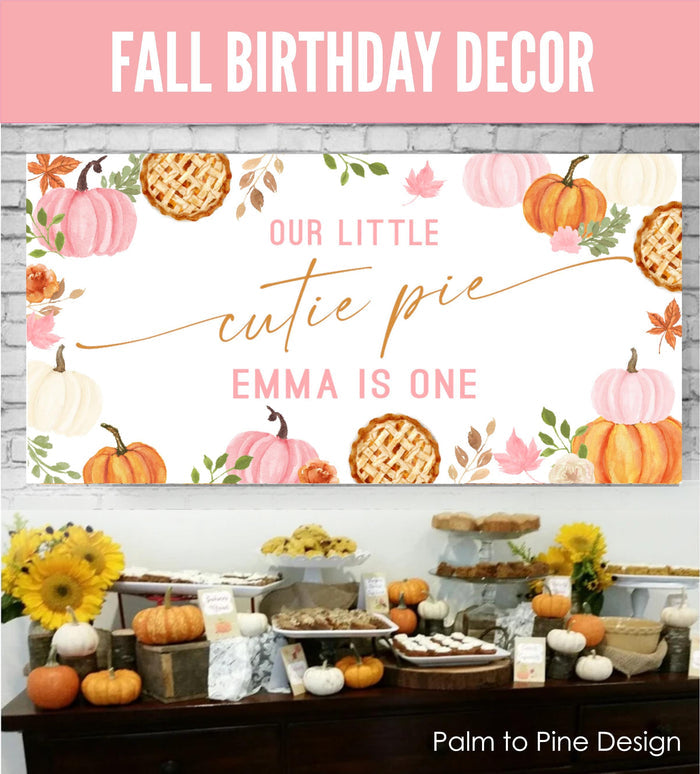 Fall-themed first birthday banner featuring pumpkins, pies, and autumn leaves in pink and orange watercolor style.  Text reads: 'Our Little Cutie Pie Emma Is One'.  A matching dessert table displays various fall-themed treats, including pies, cupcakes, and cookies.  Pumpkins and sunflowers decorate the table. The banner and table setting are perfect for a first birthday party with a fall theme.