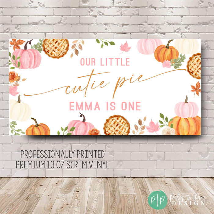 Fall-themed first birthday banner featuring pink and orange pumpkins, pie slices, and autumn leaves. The banner text reads, 'Our Little Cutie Pie Emma Is One.'  Professionally printed on premium 13 oz scrim vinyl.  Perfect for a first birthday party.