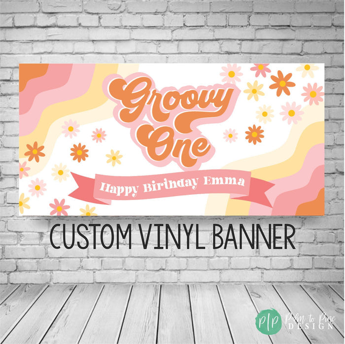 A custom vinyl banner with a groovy one theme. The banner is white with pink and orange wavy lines and yellow and white flowers. The text on the banner reads 'Groovy One' in a retro font with a pink outline. Below the text is a pink ribbon with the words 'Happy Birthday Name'