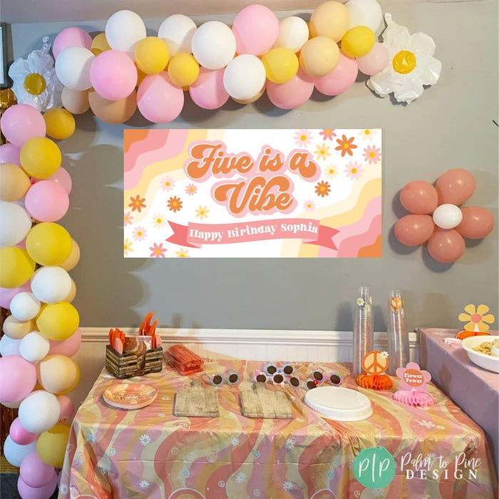 A pink and orange birthday banner with the text 'Five is a Vibe' and 'Happy Birthday Sophia'. The banner is decorated with white and yellow flowers. The banner is hanging on a wall with a garland of pink, yellow, and white balloons.