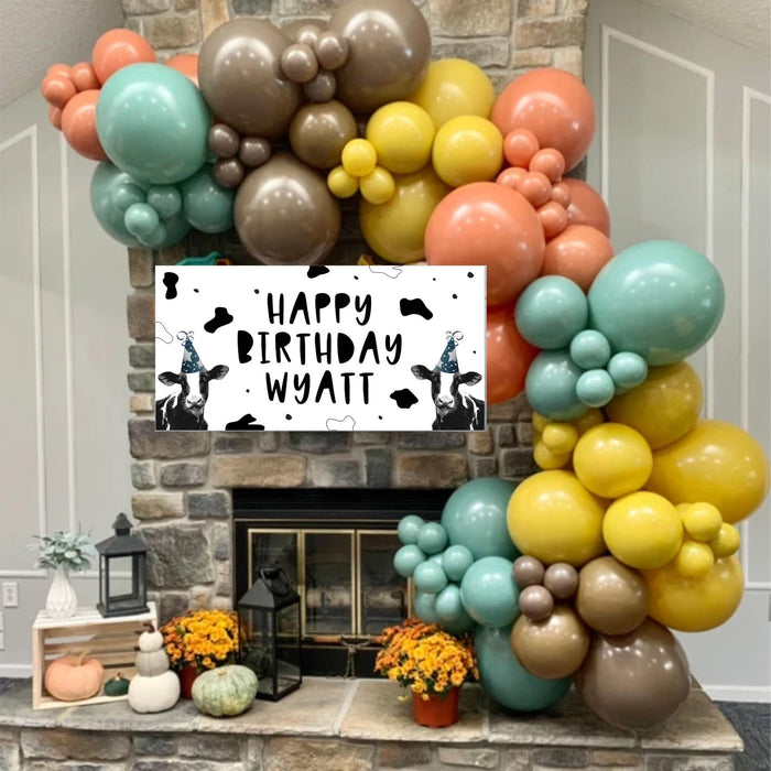 Cow Birthday Banner featuring cows in party hats with cow print background and balloon garland in front of fireplace