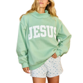 A woman wearing an oversized mint green sweatshirt with the word 'JESUS' printed in large white block letters across the chest, paired with white shorts featuring a small blue floral pattern, set against an outdoor patio backdrop.