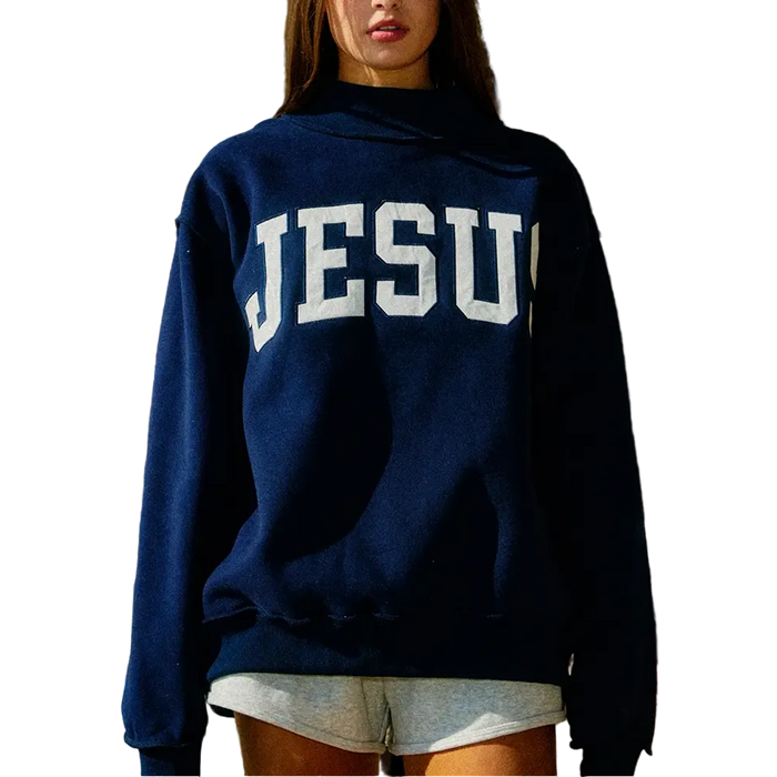A woman wearing a navy blue pullover hoodie with 'JESUS' printed in large white block letters across the chest, paired with light gray shorts, set against a rustic wooden background.