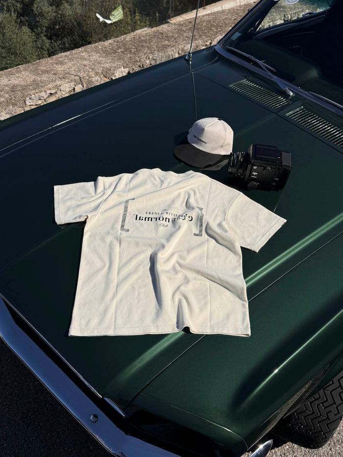 The Le T-Shirt in cream, laid flat on the hood of a dark green car. The image shows the back of the shirt with the black FRC edition mirror logo and a c'est normal cap next to a camera.