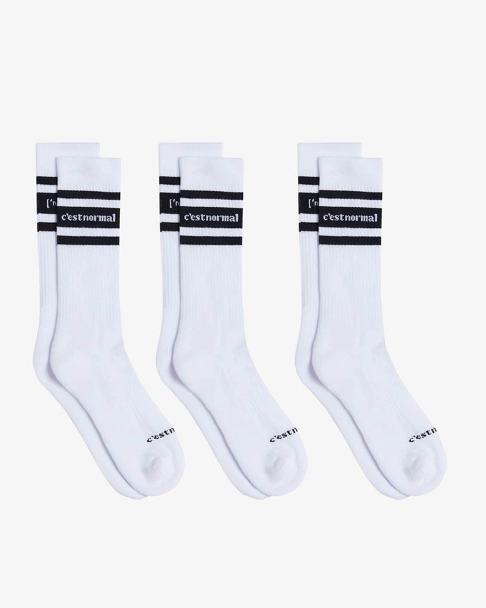 Three pairs of Les Socks in white, laid flat on a white background. The image highlights the mid-calf design with black stripes and the c'est normal logo in contrast stitching.
