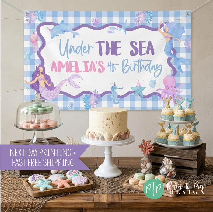 Under the sea mermaid banner displayed behind a birthday dessert table with ocean theme seashells and coral with cake