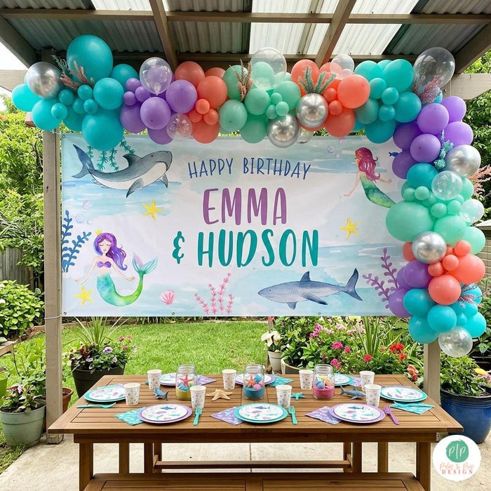 Personalized mermaid and shark birthday banner with watercolor ocean creatures hung outside in gazebo with balloon garland and under the sea table setting