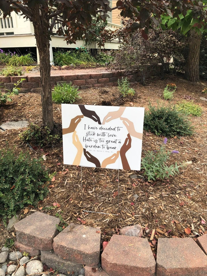 a human rights yard sign with a MLK quote about love and unity, surrounded by a garden with trees and shrubs.