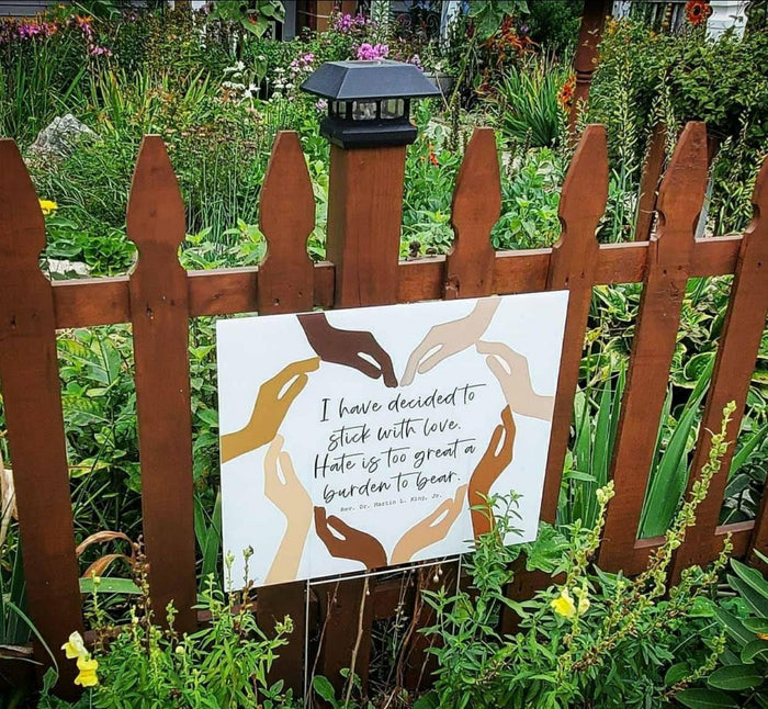 a wooden fence with a human rights yard sign attached to it, surrounded by a lush garden with various plants and flowers.