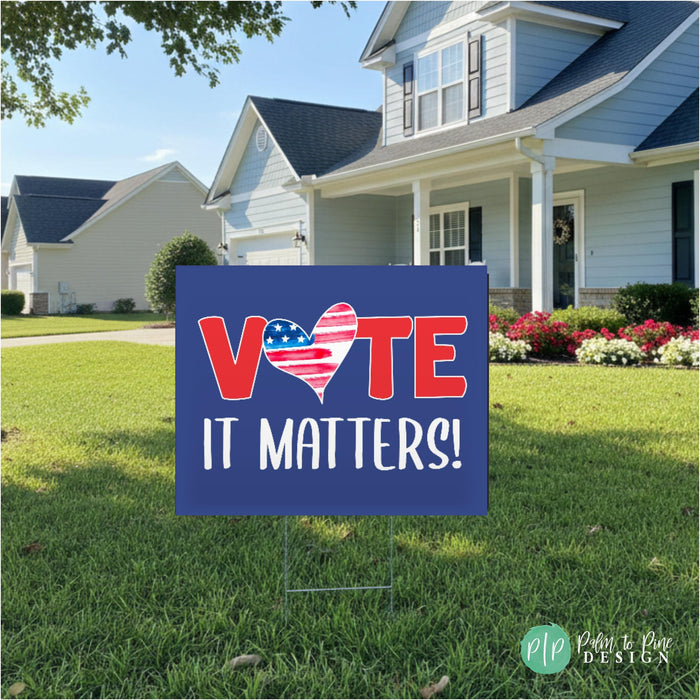 A blue sign with the text "VOTE IT MATTERS!" in red and white letters, standing in a grassy yard in front of a house.