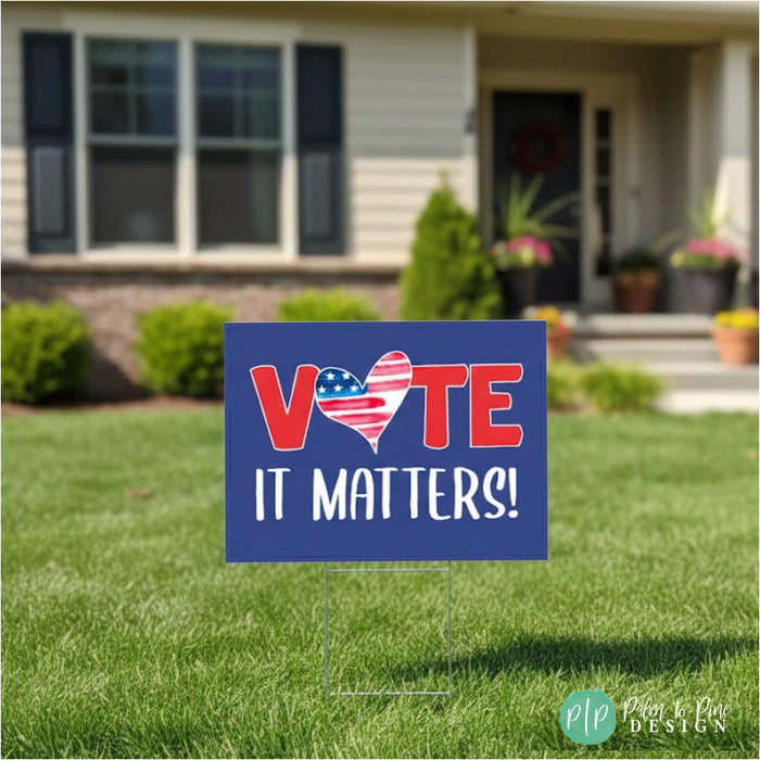 A blue sign with the text "VOTE IT MATTERS!" in red and white letters, placed on a lawn in front of a house.
