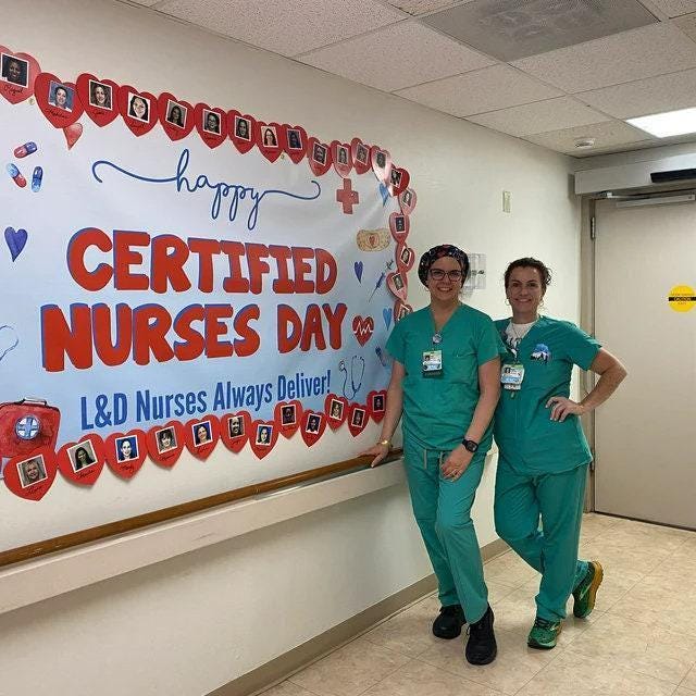 Two nurses in green scrubs stand in front of a white wall with a red and blue sign that says 'Happy Certified Nurses Day! L&D Nurses Always Deliver!' The sign is decorated with red hearts and photos of people. The nurses are smiling and looking at the camera.
