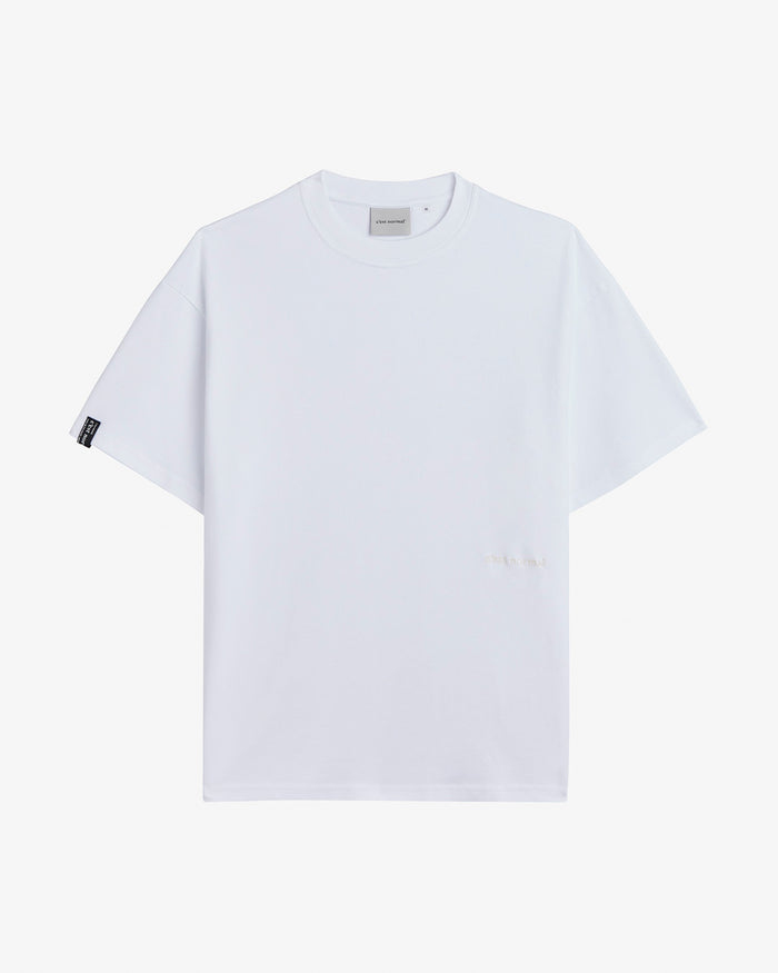 A front view of the Oversized Tonal T-Shirt in white, laid flat on a white background. The image highlights the classic ribbed collar, the tonal logo on the lower front, and the black c'est normal tag on the right sleeve.