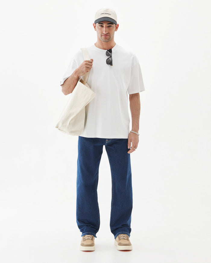 A man wearing the Oversized Tonal T-Shirt in white, paired with blue jeans, a c'est normal cap, and sunglasses tucked into the collar. He is also holding a white tote bag over his shoulder against a white background.