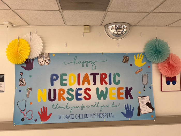 A vibrant blue banner celebrates Pediatric Nurses Week.  Rainbow-colored lettering proclaims 'Happy Pediatric Nurses Week,' expressing gratitude with 'Thank you for all you do!'  UC Davis Children's Hospital is credited below.  The banner features colorful handprints, medical icons, and playful illustrations.  Paper fans in yellow, white, teal, and pink decorate the surrounding area.
