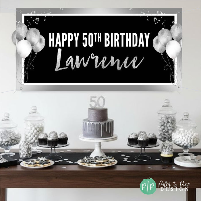 A black and silver birthday banner with the text "Happy 50th Birthday Lawrence" is displayed above a table filled with desserts, including a cake and various candies.