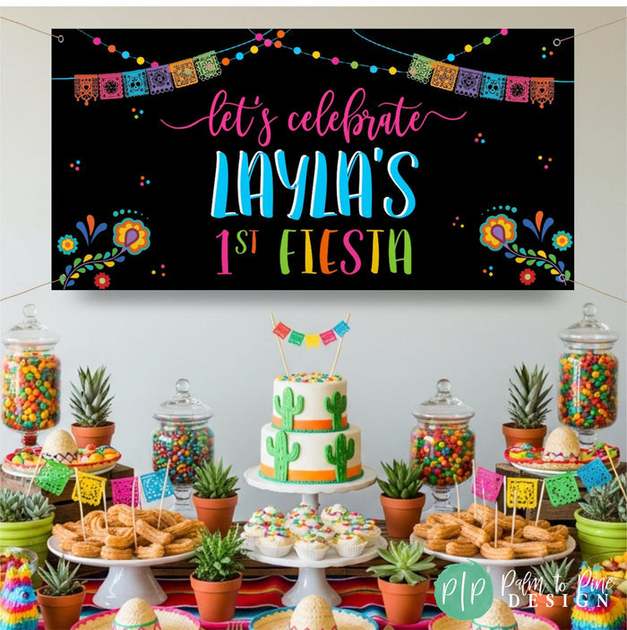A colorful themed banner with the text "Let's celebrate Layla's 1st Fiesta" is displayed above a table filled with various mexican desserts, including a cake, cupcakes, and candy jars.