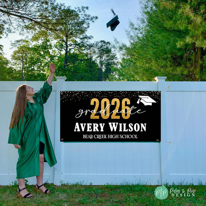 Custom Class of 2026 graduate banner hung outdoors on fence with a high school graduate in cap and gown throwing her hat