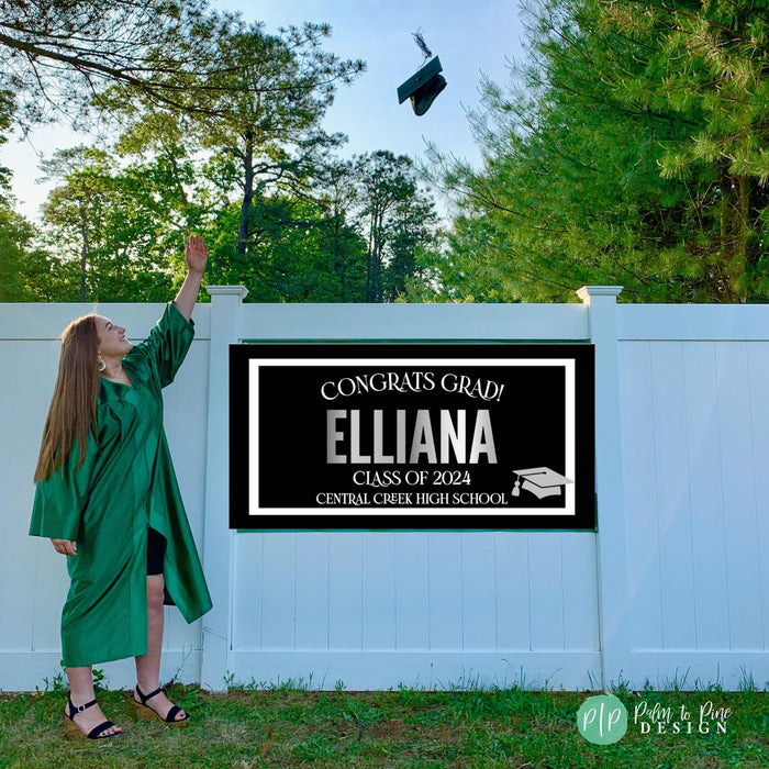 A black and white graduation sign with silver text that reads 'Congrats Grad! Elliana Class of 2025 Central Creek High School' with a graduation cap icon. The sign is displayed on a white picket fence.