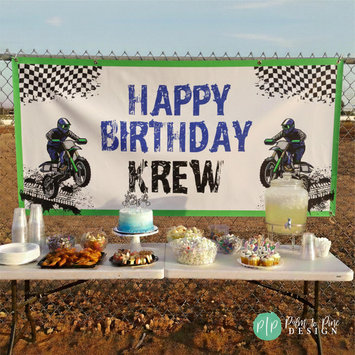 Outdoor dirt bike race track birthday celebration featuring a personalized birthday banner hanging from a chain link fence and attached with grommets. Below the banner is a folding table with potluck style party desserts