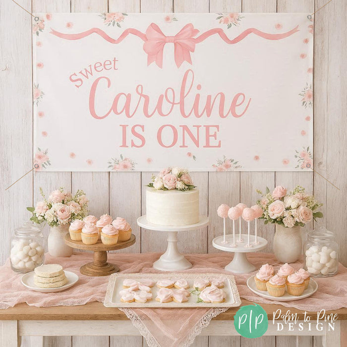 Girls first birthday banner in bow and flower theme, hung behind a feminine pink dessert table