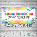 A colorful science-themed birthday banner with the text 'Happy Birthday Carson's Science Lab' and the words 'Custom Vinyl Banner' at the bottom. The banner has a watercolor background with a variety of science-related images, including beakers, test tubes, molecules, and a microscope.