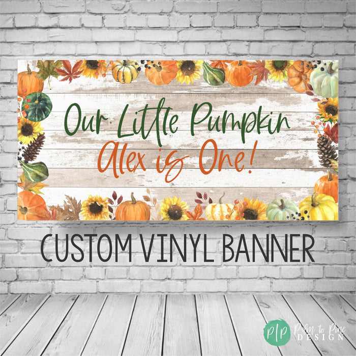 A custom vinyl banner with a rustic wood background and fall-themed decorations. The banner reads 'Our Little Pumpkin Alex is One!' with sunflowers, pumpkins, and fall leaves. The text 'CUSTOM VINYL BANNER' is printed below the design.