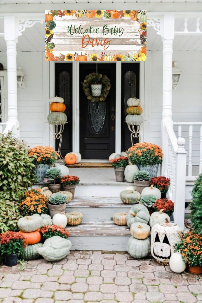 A white house with a wooden front porch decorated with a welcome baby banner with fall foliage and pumpkins. The banner reads 'Welcome Baby Davis'. The porch is decorated with pumpkins and fall flowers in pots.
