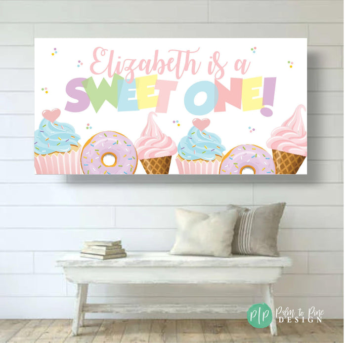 First birthday banner in pastel rainbow hues with charming dessert illustrations—cupcakes, donuts, and ice cream cones for a sweet-themed party.