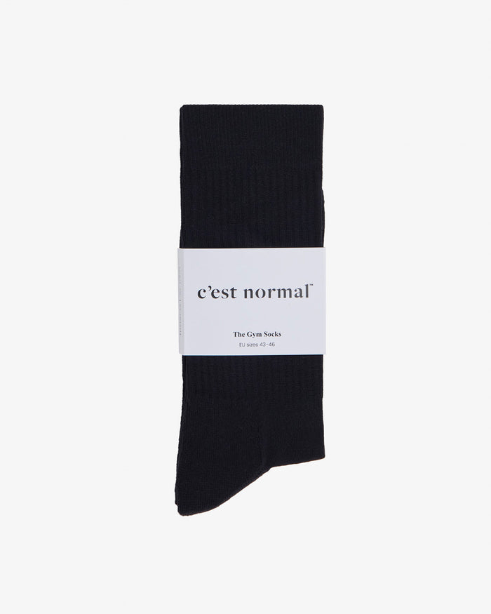 A pair of black The Athlétique Socks laid flat on a white background, secured with a white paper band featuring the c'est normal logo.