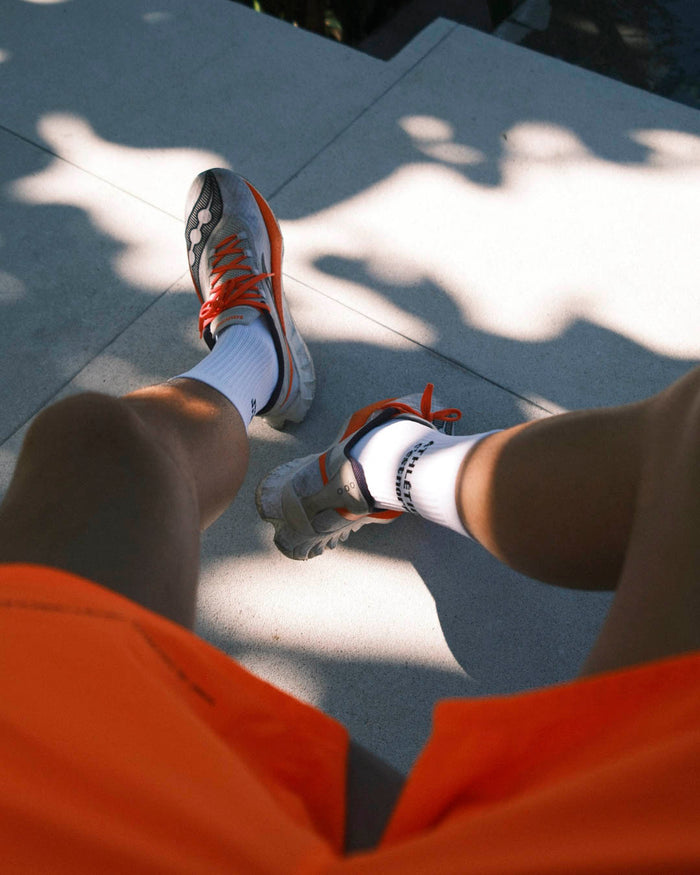 A first-person view of a person wearing the white Athlétique Socks, orange shorts, and running shoes. The image highlights the black c'est normal logo on the side of the right sock.