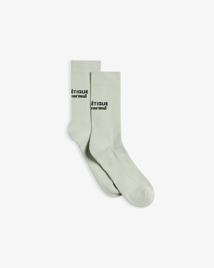 A pair of The Athlétique Socks in sage green, laid flat and slightly overlapping on a white background. The image highlights the ribbed cuffs and the black embroidered c'est normal logo at mid-calf.