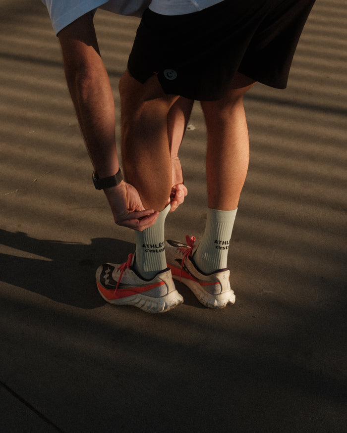 A person wearing The Athlétique Socks in sage green, paired with running shoes and black shorts, bends down to adjust them. The image highlights the mid-calf length and the black ATHLÉTIQUE and c'est normal text printed on the side.