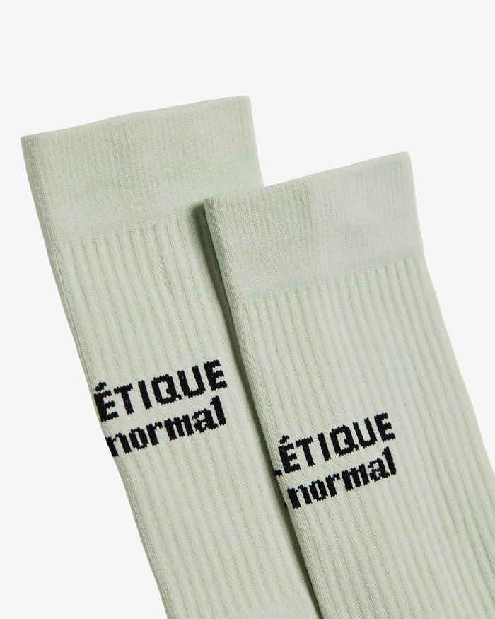 A close-up view of The Athlétique Socks in sage green, showing the ribbed texture and the black contrast stitching of the c'est normal logo at mid-calf.