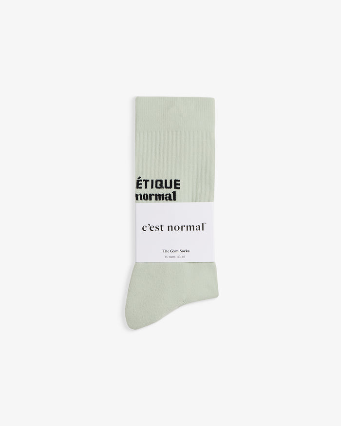 The Athlétique Socks in sage green, folded and laid flat on a white background. The image highlights the ribbed mid-calf with contrasting black stitched text and a white c'est normal branded sleeve.