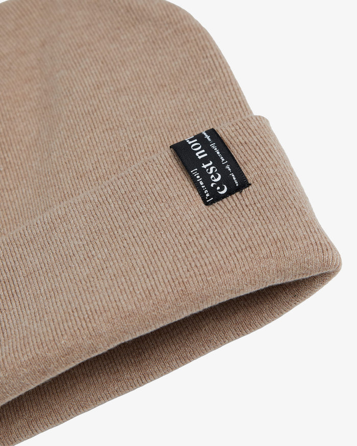 A close-up shot of the soft, fine knit of The Beanie in beige, highlighting the fold-over cuff and the signature black c'est normal label.