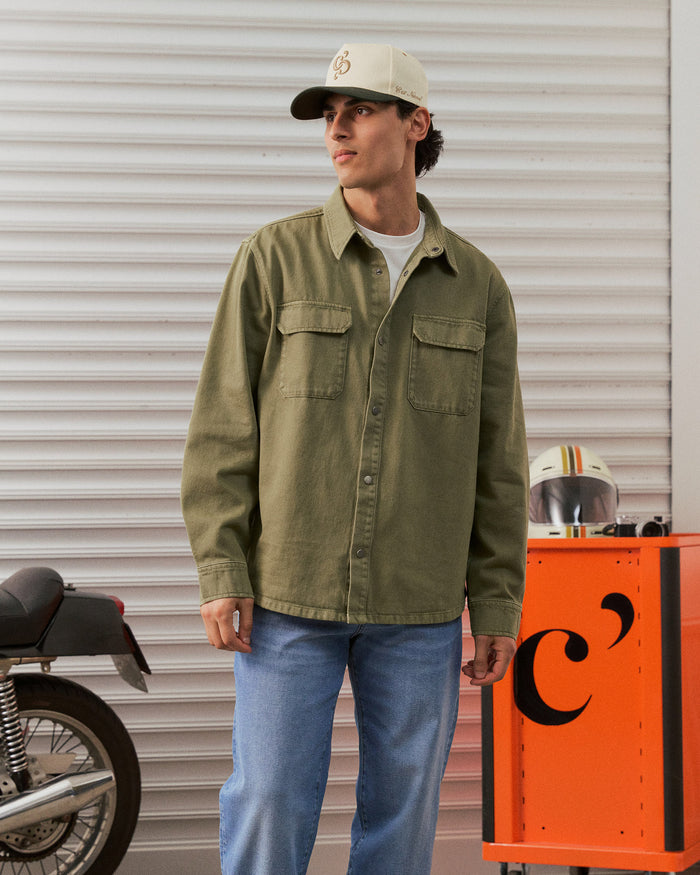 The Bilbao Overshirt in olive — styled with blue jeans and a cap next to a motorcycle