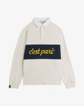 A front view of The c'est pari Rugby Shirt in cream, laid flat on a white background. The image highlights the white collar, covered placket, and the dark blue horizontal stripe across the chest featuring the yellow c'est pari logo. A black c'est normal tag is visible on the right cuff.