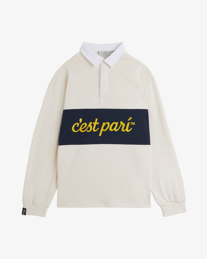 A front view of The c'est pari Rugby Shirt in cream, laid flat on a white background. The image highlights the white collar, covered placket, and the dark blue horizontal stripe across the chest featuring the yellow c'est pari logo. A black c'est normal tag is visible on the right cuff.