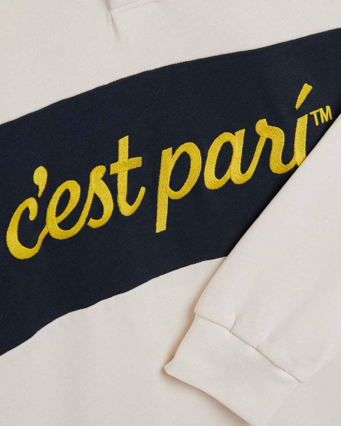 A detailed close-up of The c'est pari Rugby Shirt in cream, highlighting the yellow embroidered c'est pari logo across a wide navy blue horizontal stripe.