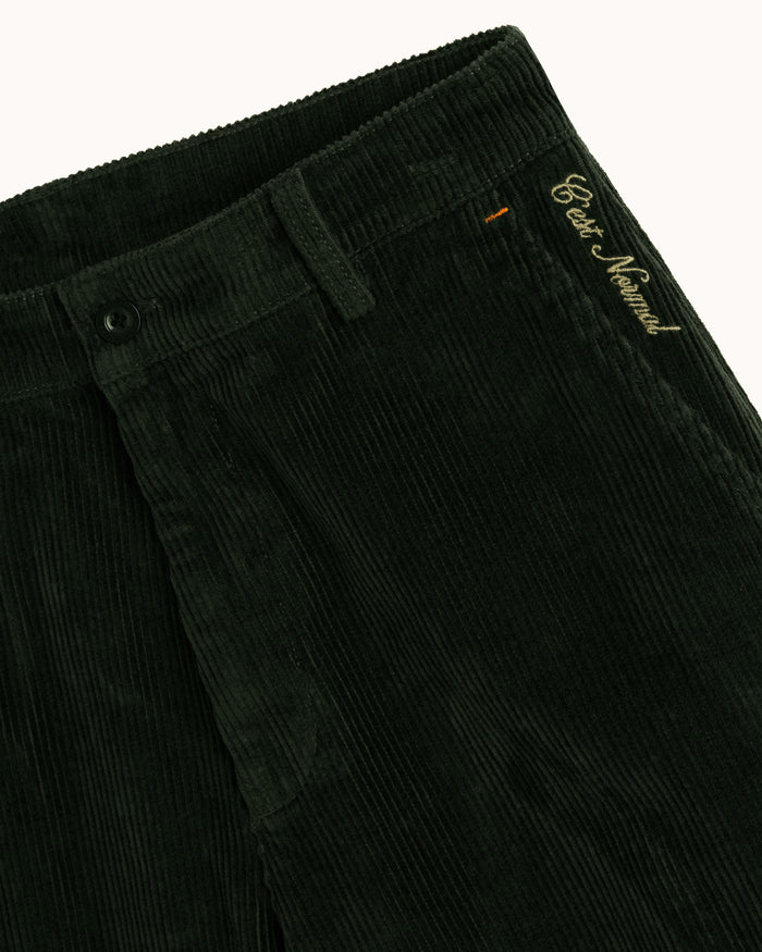 A detailed close-up of the top front of The CÔTELÉ Corduroy Straight Pants in forest green, laid flat on a white background. The image highlights the corduroy texture, waistband with belt loops, button closure, and the embroidered c'est normal logo at the left hip.