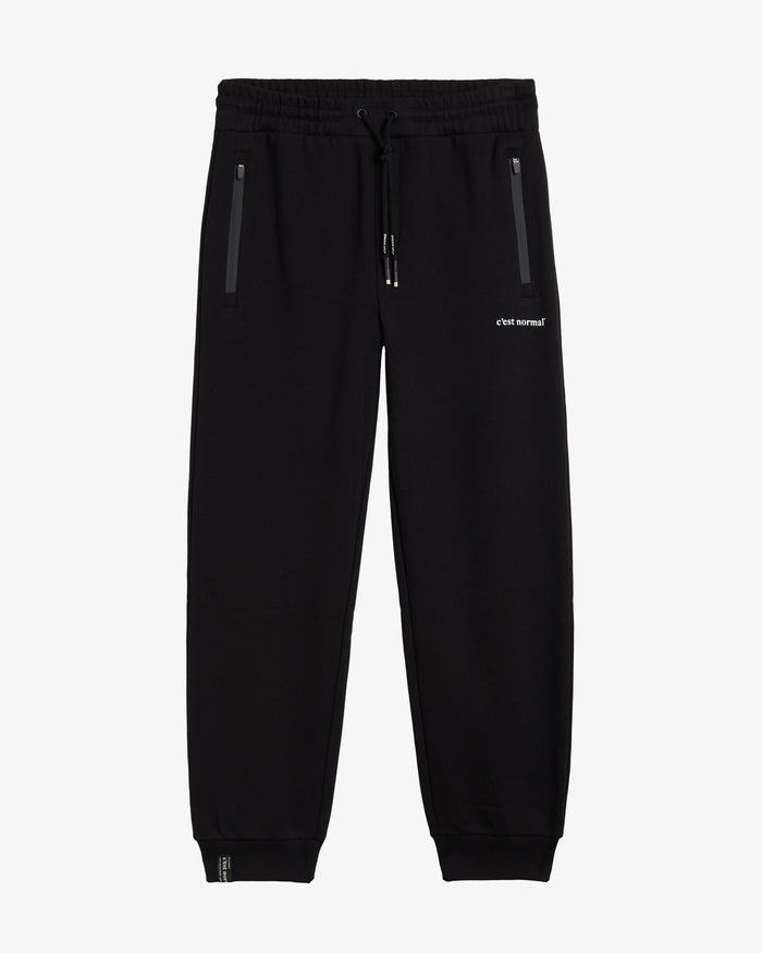 A front view of The Gym Pants in black, laid flat on a white background. The image highlights the elastic waist with a drawstring, hip pockets with premium taped zippers, and the white c'est normal logo on the left thigh.