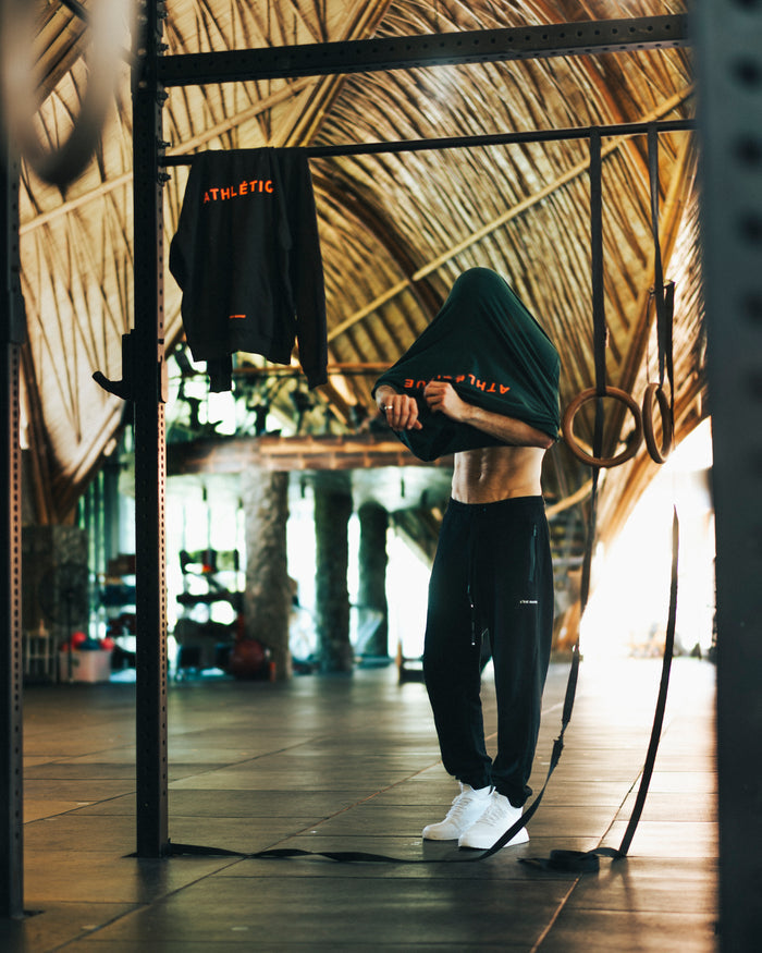A man in a gym pulling a dark t-shirt over his head, wearing The Gym Pants in black and white sneakers. The image showcases the pants' relaxed fit, zippered pockets, and the c'est normal logo on the left thigh. A dark hoodie hangs on a barbell in the foreground.
